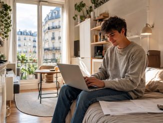 Jeune alternant assis sur son lit dans son studio parisien, en train de monter son dossier logement sur son ordinateur portable